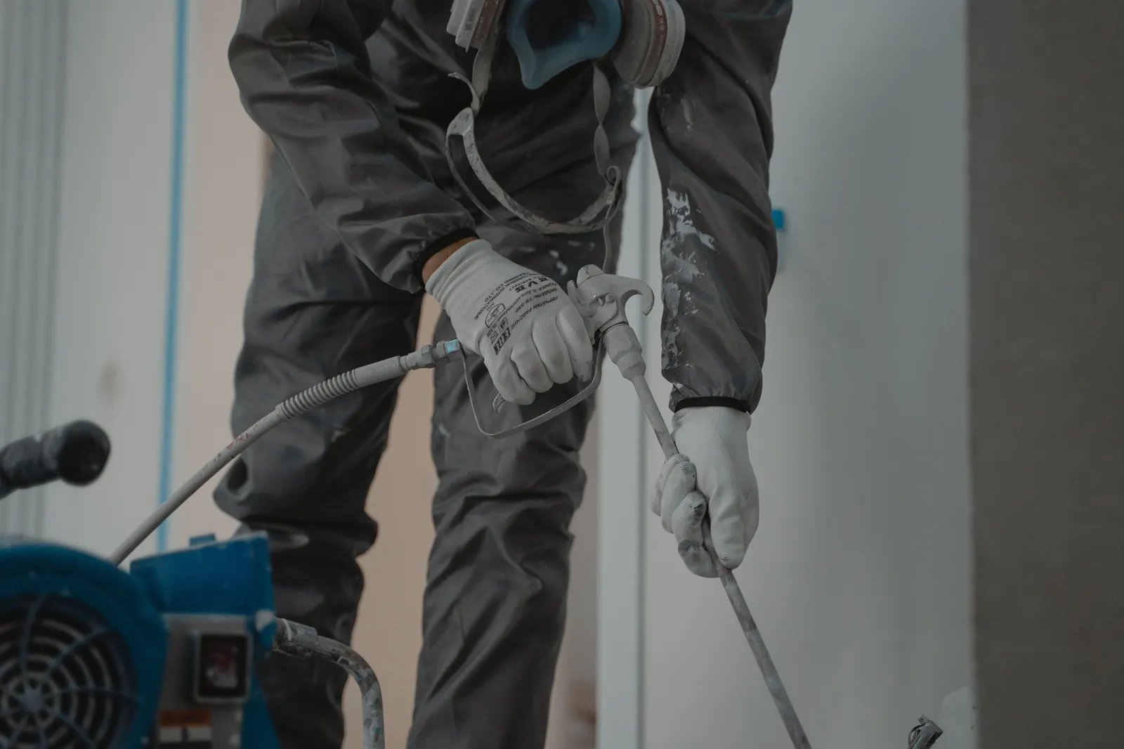 An airless paint sprayer and spray gun Close-up of gloved hands holding an airless spray gun and extension wand, with a blue paint sprayer in the foreground