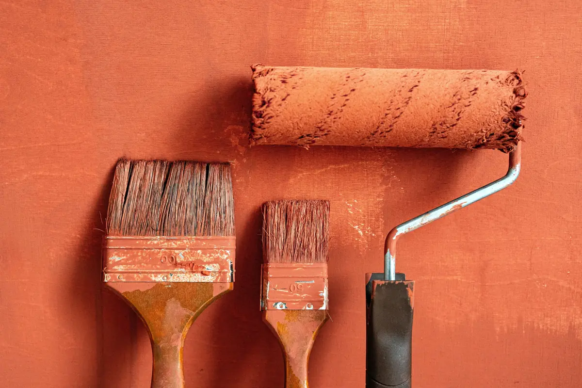 Two paint brushes and a roller resting against a freshly painted terracotta wall