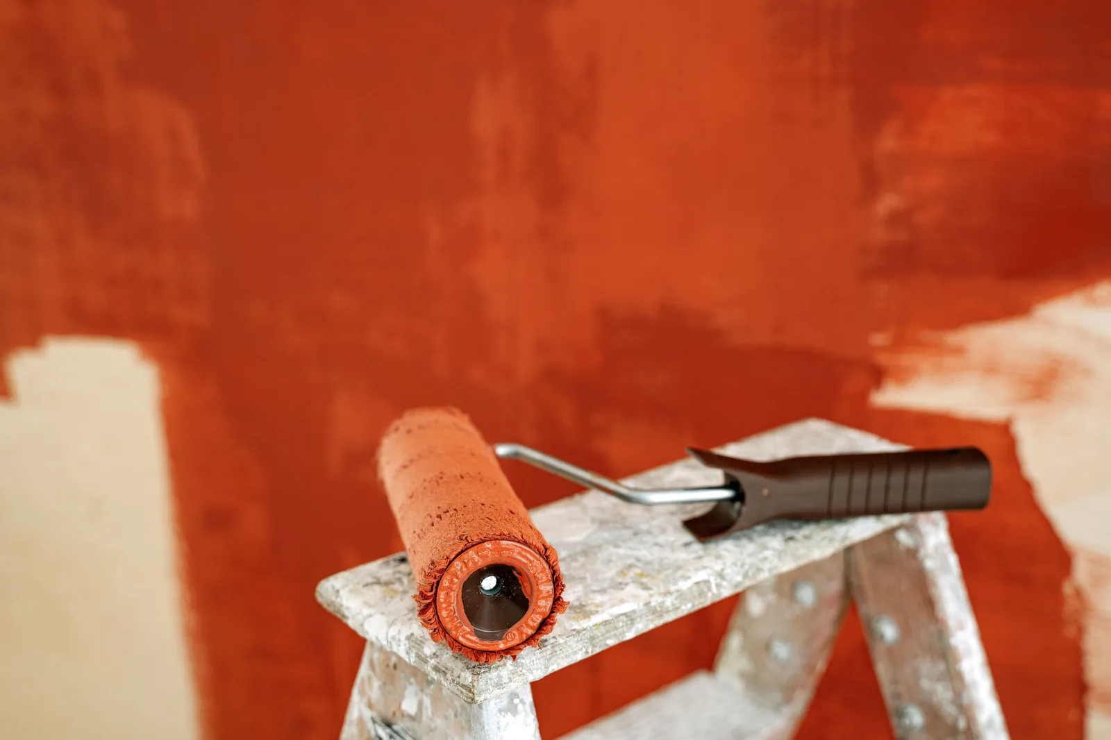 Paint roller with an orange cover resting on a paint-spattered stepladder in front of a partly painted terracotta wall