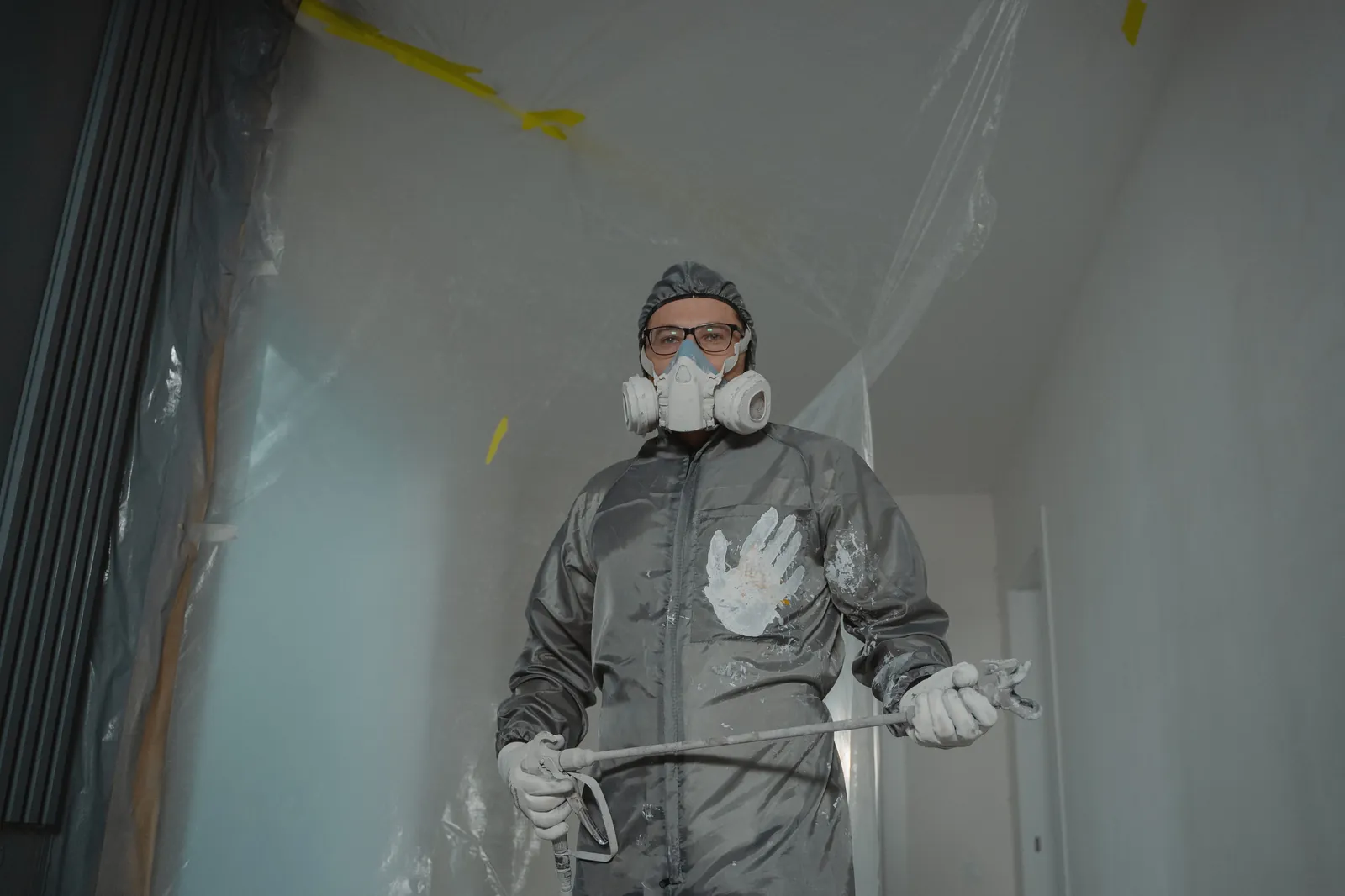 Painter in paint-spattered coveralls, a half-face respirator and safety glasses holding a spray gun in a room masked with plastic sheeting