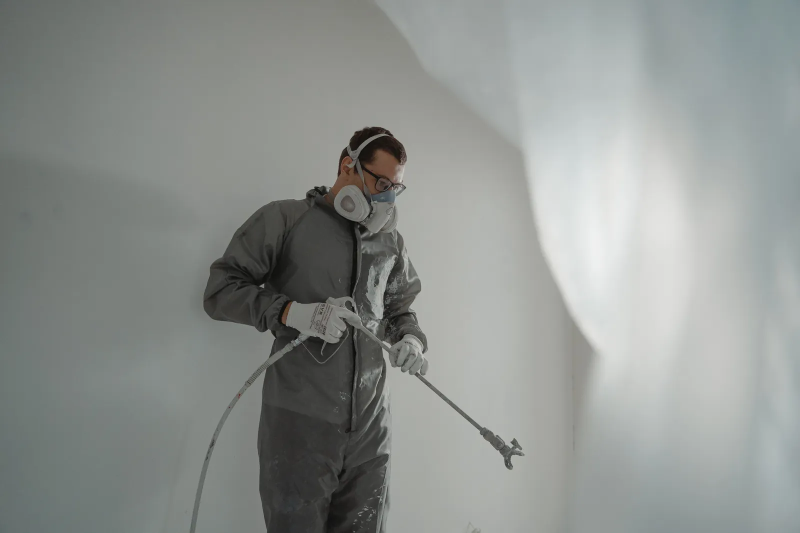 Painter in grey coveralls, a respirator and safety glasses holding a spray gun with an extension wand in front of a white wall