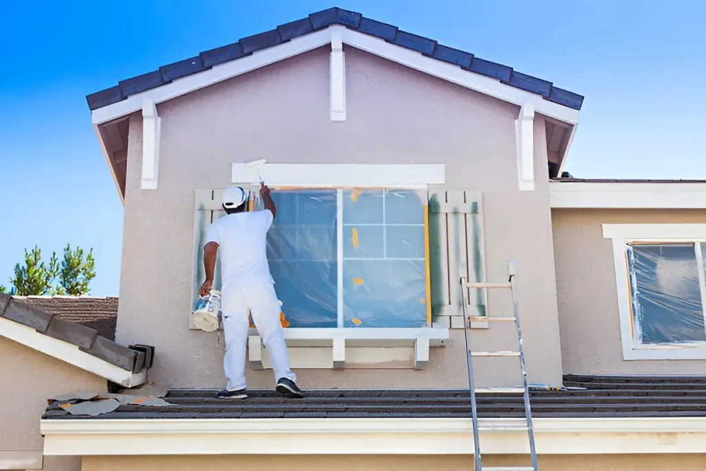 Painter on a roof painting the trim around a second-floor window, with the glass and shutters masked off